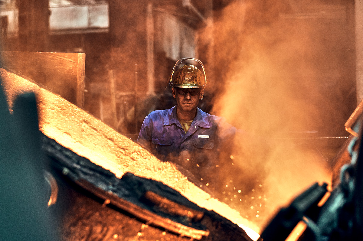 Gießer mit Schutzkleidung hinter großem Kessel mit glühendem Kupfer