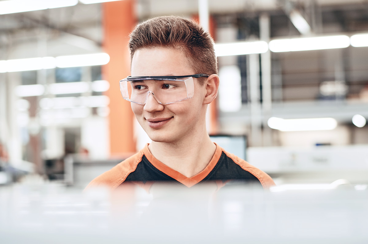 Lächelnder, junger Auszubildender trägt Arbeitskleidung und Schutzbrille