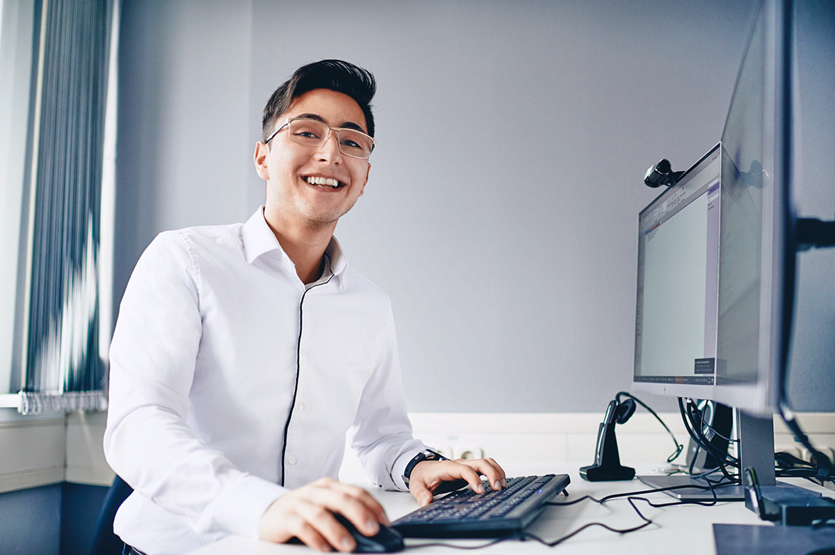 Junger Auszubildender aus der IT steht vor einem Computer und lächelt in die Kamera